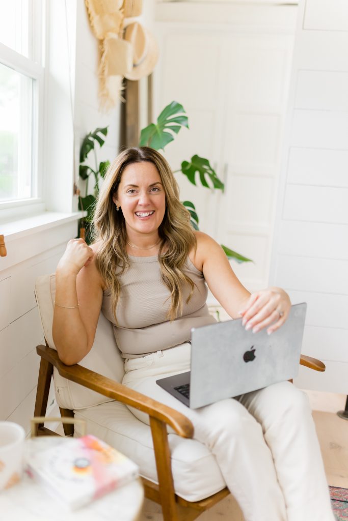 A photo of Kate Lombardo holding her open laptop on her lap, smiling at the camera in a white chair for the Business of Yoga Teacher Training.