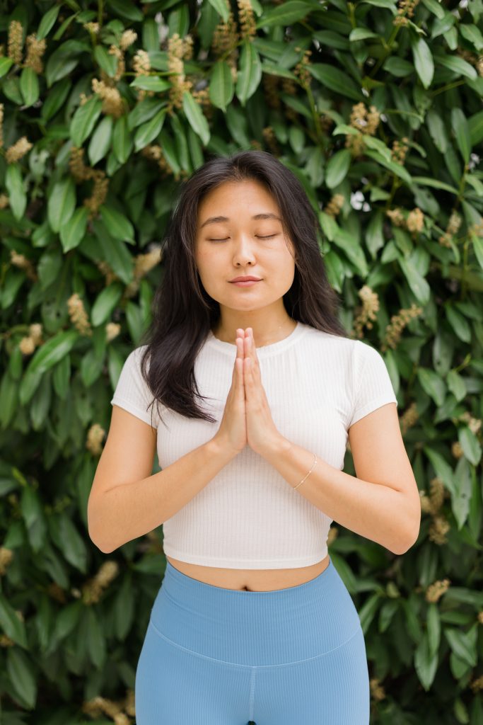 Dari, a YogaRenew student, with her eyes closed and hands in prayer at the center of her chest in front of green shrubs.