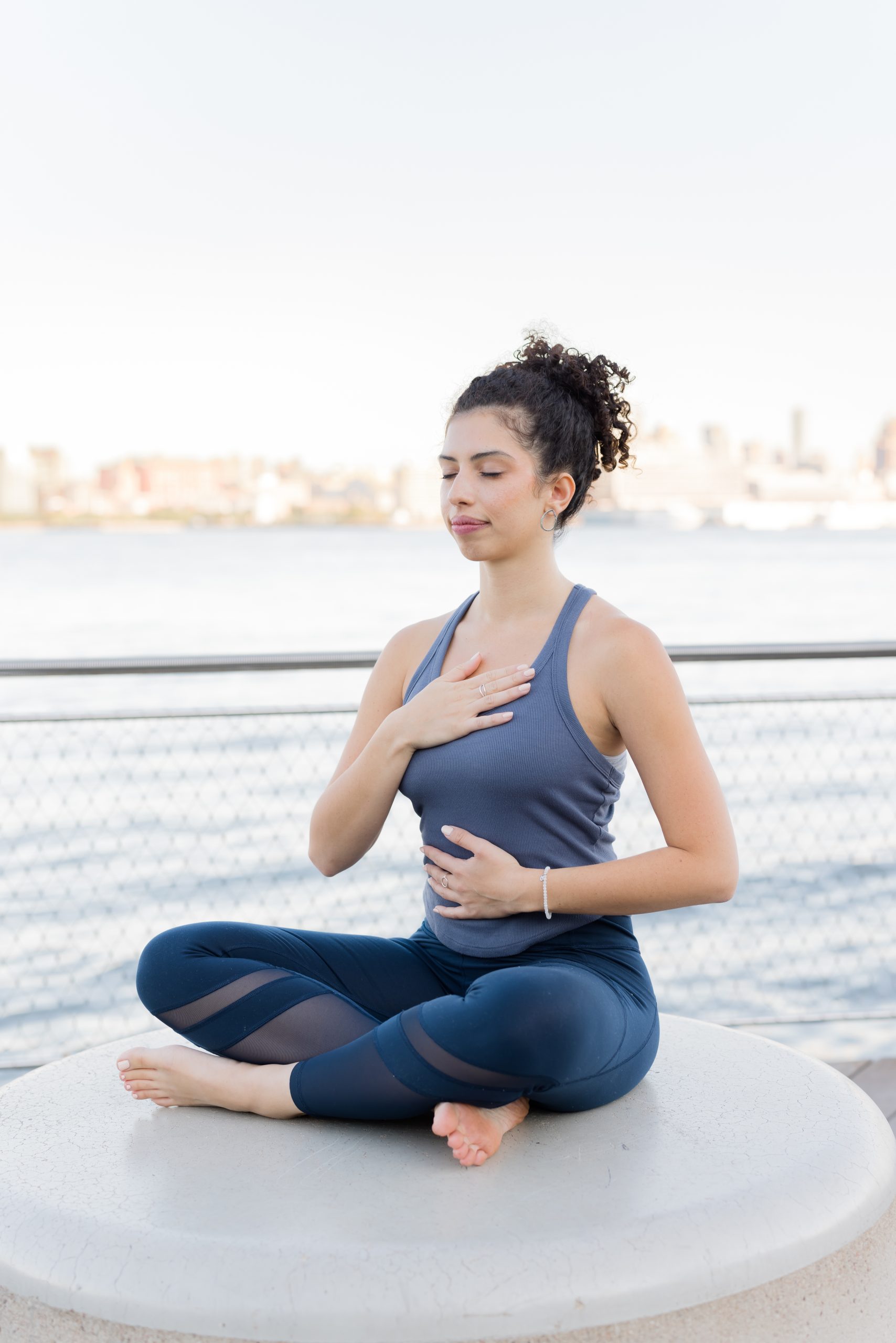 Lindsay Monal sits at Hoboken Skyline with hands over heart meditating and taking deep breaths