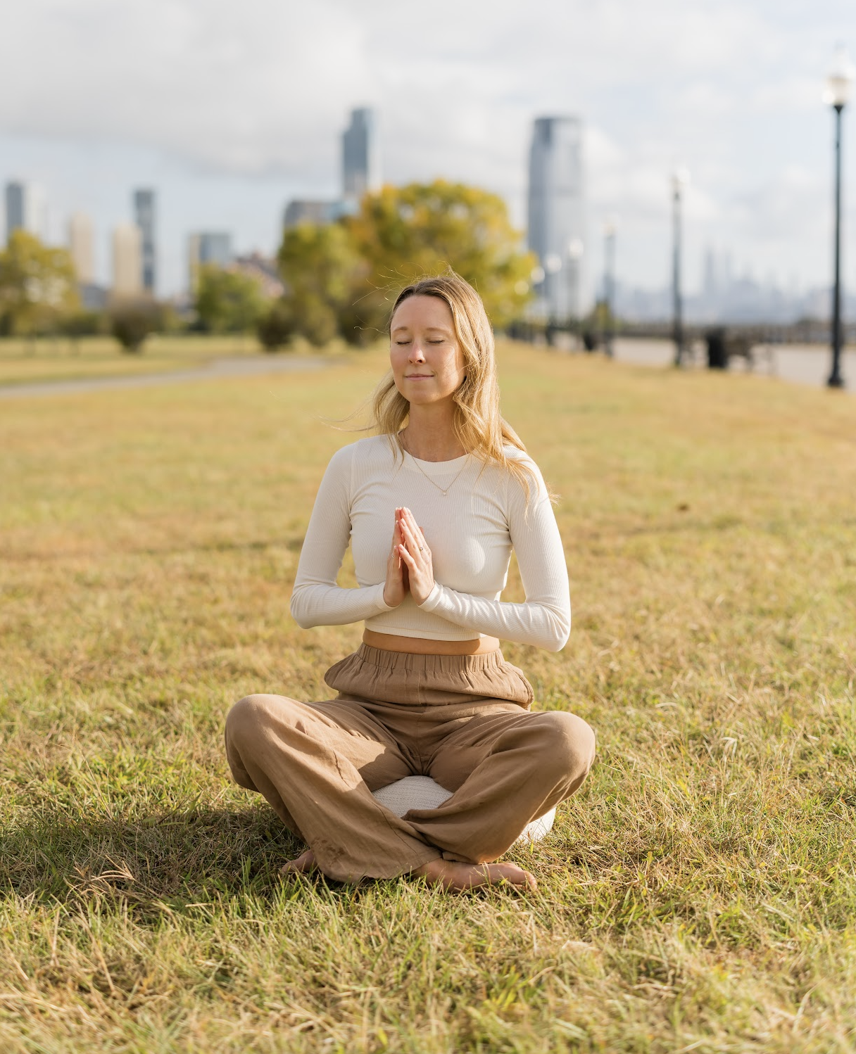 Kate Wall sitting in a field of grass in a park in Hoboken, NJ meditating with the NYC skyline behind her for her Reiki Level 1 Certification training